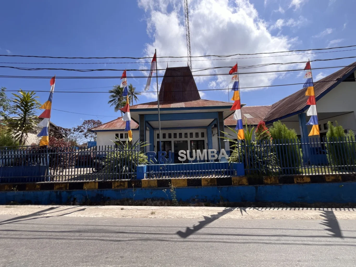 The white and blue RRI Sumba building with a traditional roof stands behind a blue fence adorned with Indonesian and colorful flags under a bright blue sky.