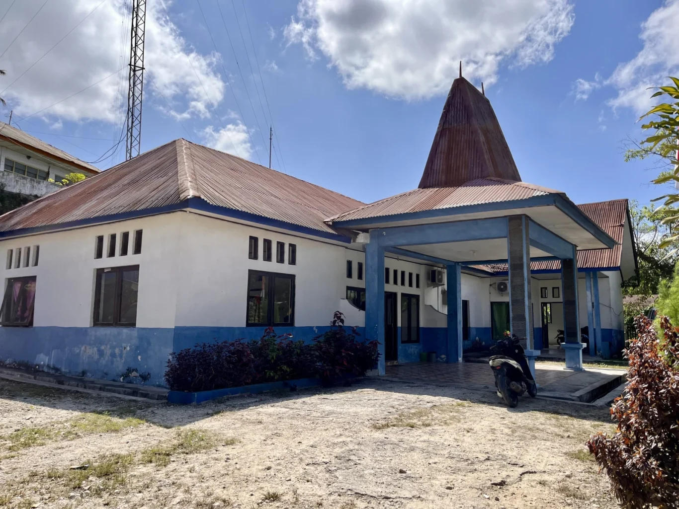 The white and blue Radio Republik Indonesia Sumba building, with a traditional conical roof and reddish-brown corrugated metal, stands under a sunny sky with a radio tower visible.