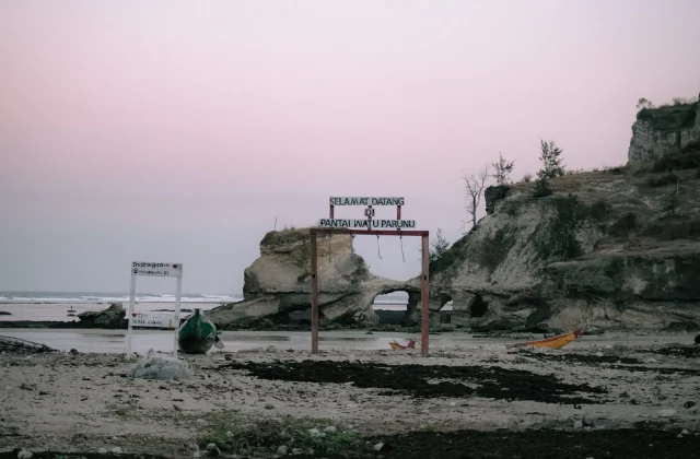 Watuparunu Beach features a welcome sign and a natural rock arch, with two small boats on the sandy shore under a soft pink sky, flanked by rocky cliffs.