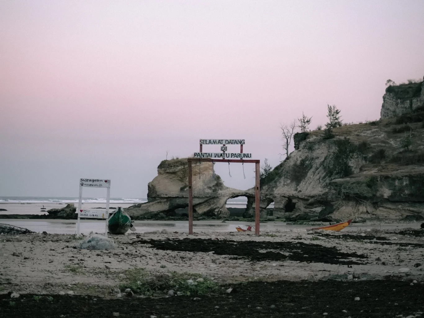 Watuparunu Beach features a welcome sign and a natural rock arch, with two small boats on the sandy shore under a soft pink sky, flanked by rocky cliffs.