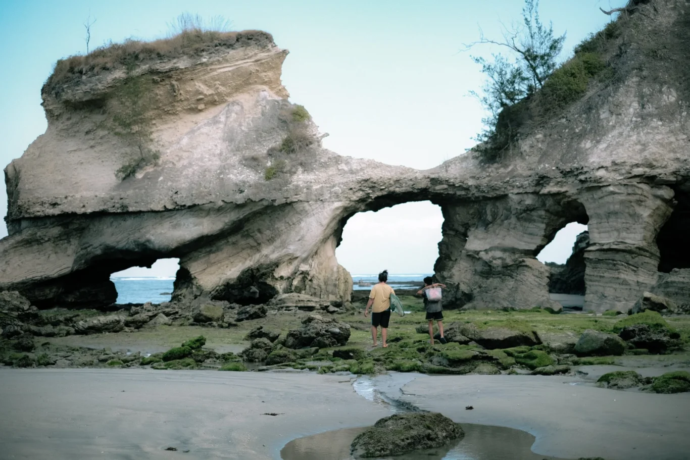 Two people walk on a sandy, mossy beach towards the ocean, passing under large, light-colored rock formations with natural arches.