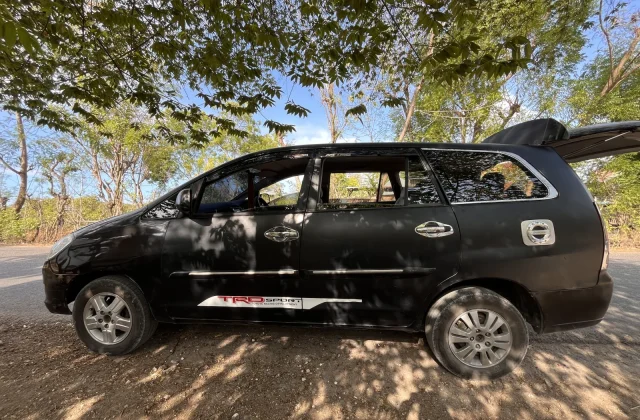 A black Toyota Innova car with an open trunk is parked on a dirt road under green trees, with sunlight casting shadows.