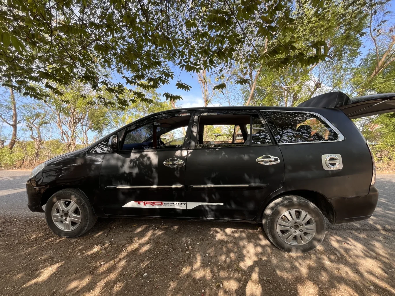 A black Toyota Innova car with an open trunk is parked on a dirt road under green trees, with sunlight casting shadows.