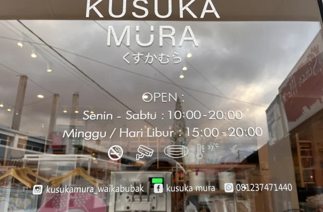 The glass storefront of Kusuka Mura in Waikabubak displays its name, operating hours, social media details, and safety icons, with reflections of a cloudy sky.
