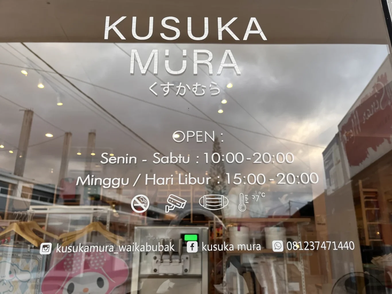 The glass storefront of Kusuka Mura in Waikabubak displays its name, operating hours, social media details, and safety icons, with reflections of a cloudy sky.