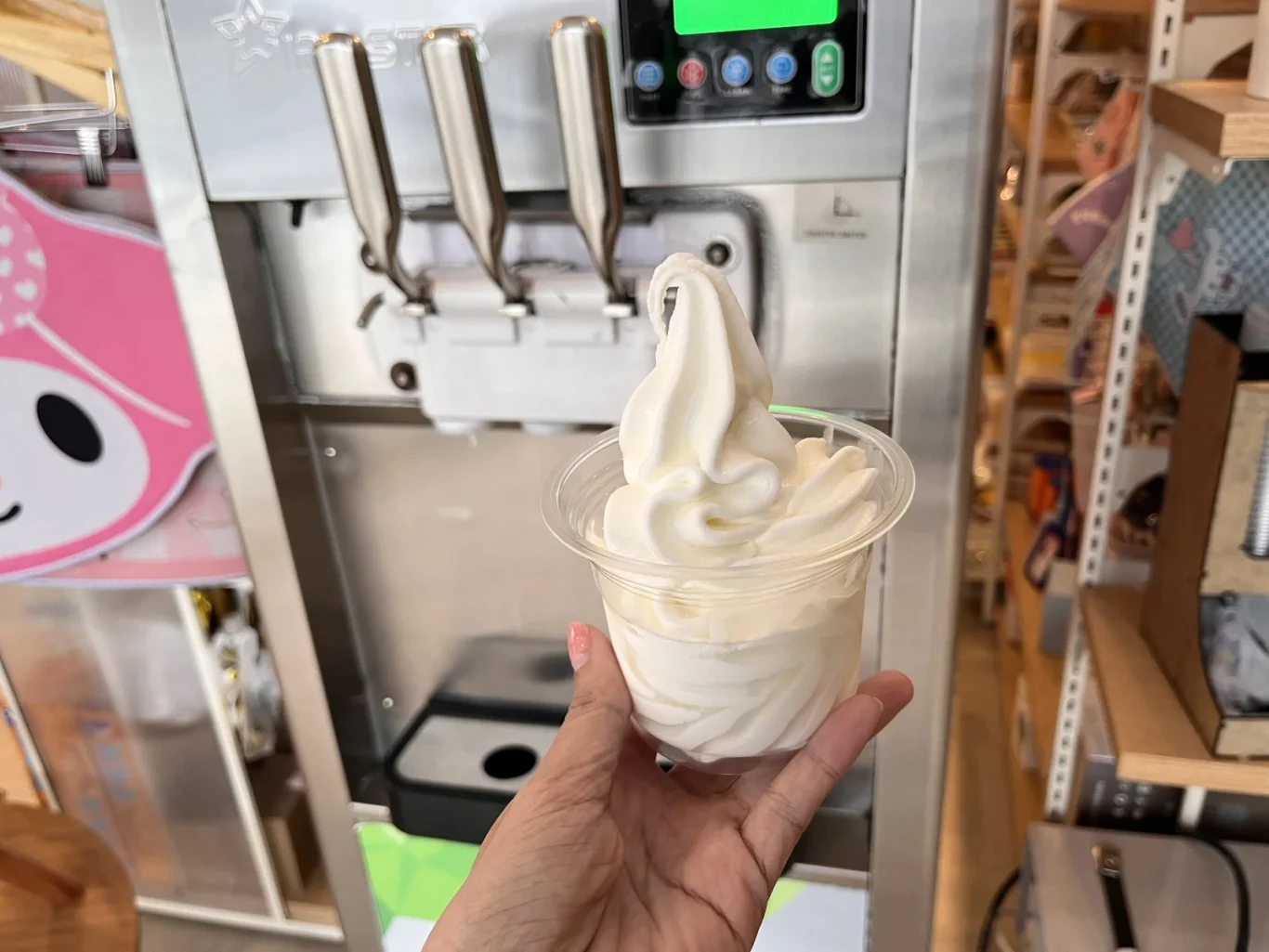 A hand holds a clear cup of white soft-serve ice cream in front of a stainless steel machine and a pink character cutout.
