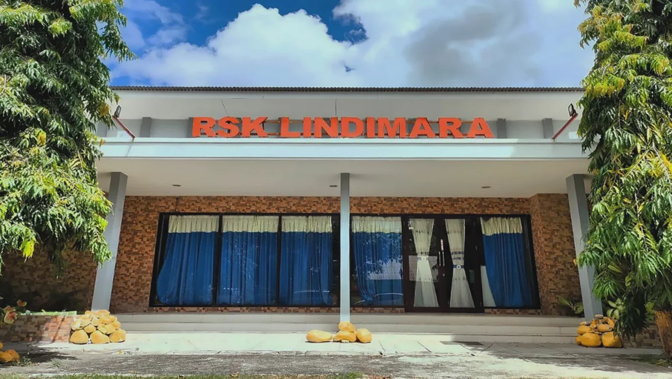 The front of a white and brown building with "RSK LINDIMARA" in orange letters, framed by two tall green trees under a cloudy blue sky.