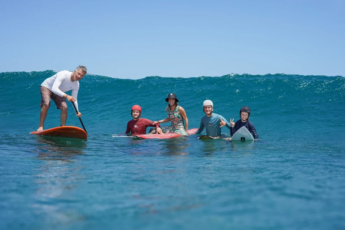 A family of five, including two adults and three children, smiles in the blue ocean with a large wave behind them; one adult paddleboards while the others are on surfboards.