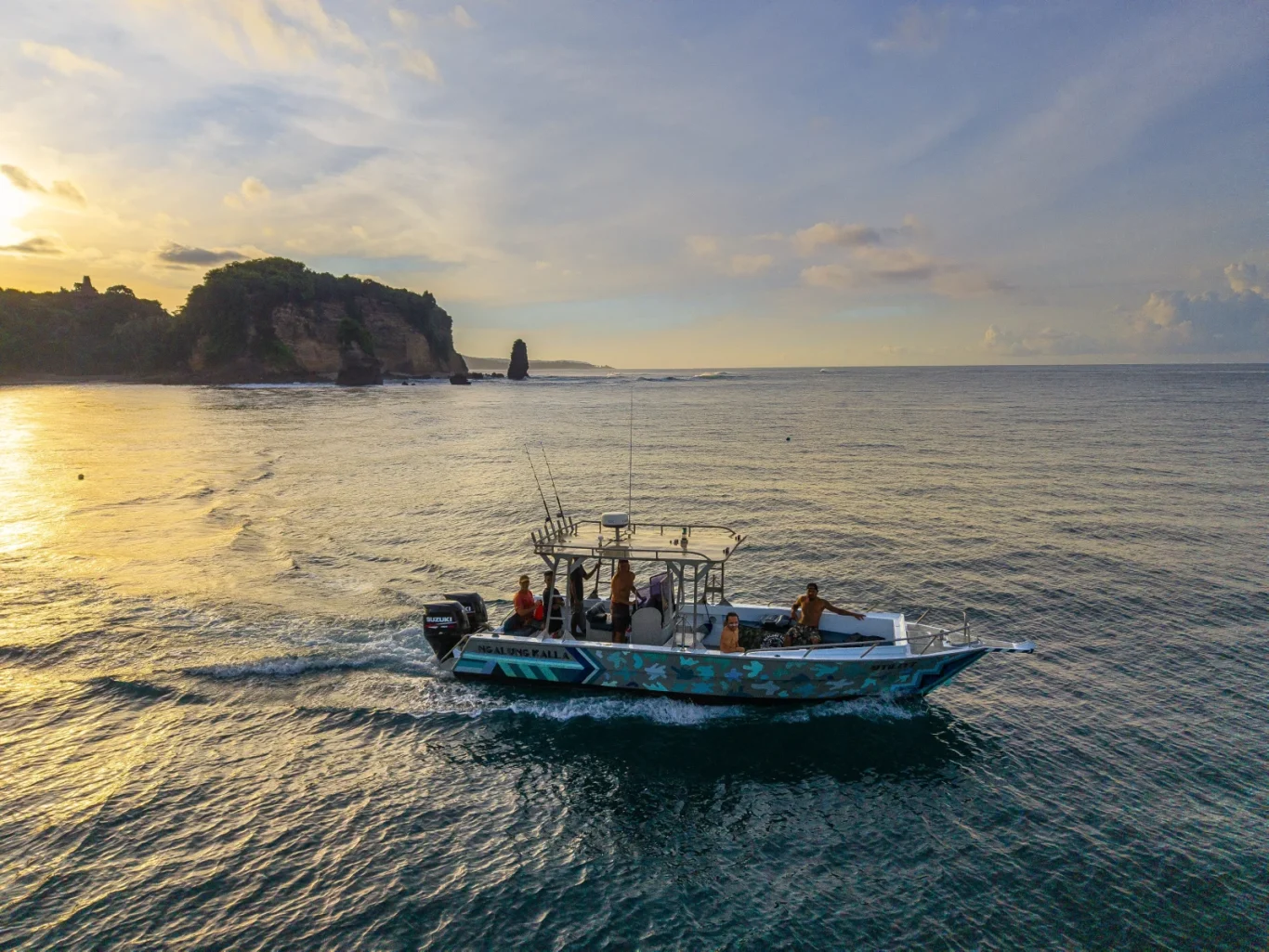 A motorboat with several people cruises on golden-hued ocean waters towards a rocky, tree-covered coastline under a sunset sky.