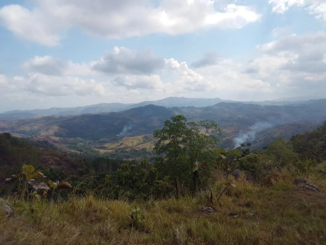 Rolling hills and distant mountains stretch under a partly cloudy blue sky, with dry grass and green foliage in the foreground. Wisps of smoke rise from several valleys.