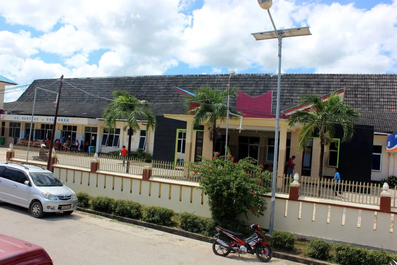 A cream-colored building with a dark roof and pink accents stands under a cloudy blue sky, with palm trees, a silver car, and a red motorcycle on the street.