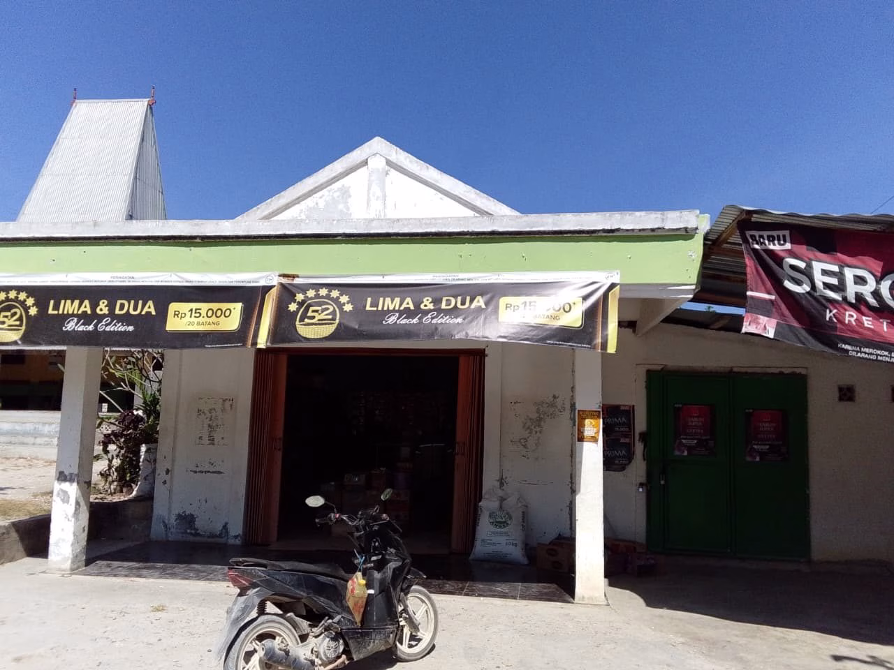 A small, white building with a green overhang features "LIMA & DUA" banners and an open entrance, with a black scooter parked outside under a clear blue sky.