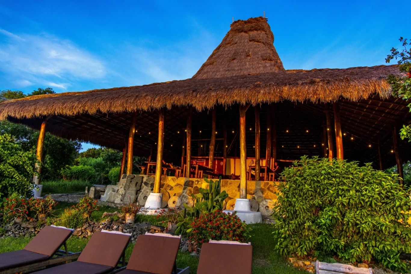 A traditional building with a conical thatched roof and open-air lower level, lit by string lights. Lush plants and lounge chairs are on grass.