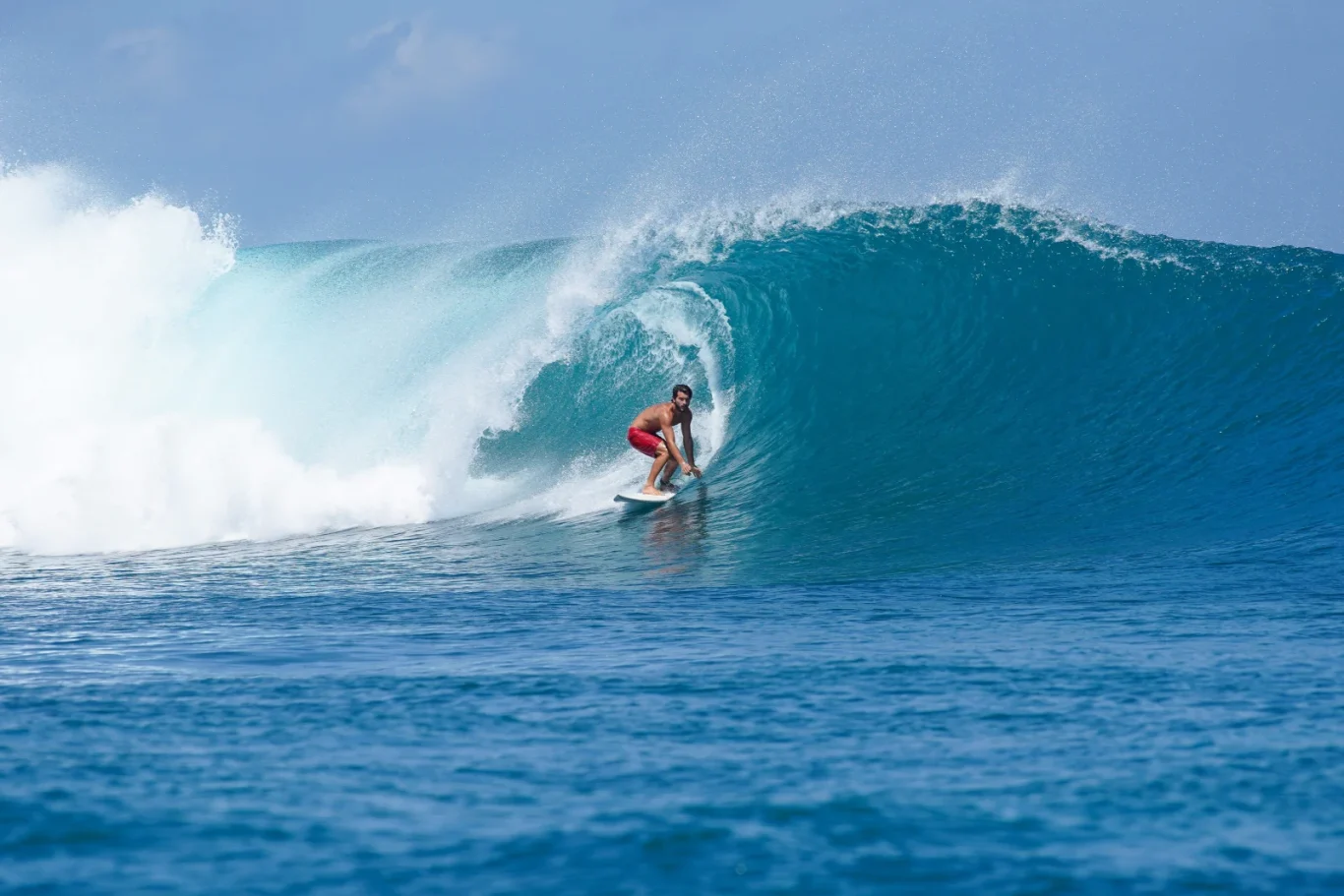 A male surfer in red trunks rides a white surfboard inside a large, barreling blue ocean wave under a bright sky.