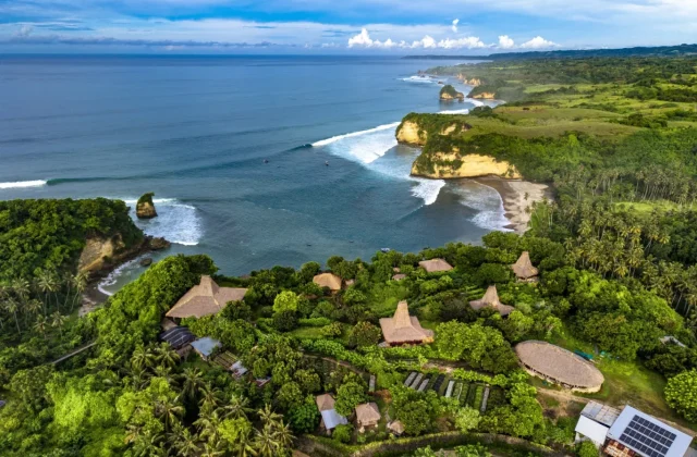 Aerial view of a vibrant green coastline with traditional thatched-roof buildings, palm trees, sandy beaches, and rocky cliffs along a deep blue ocean with white waves.
