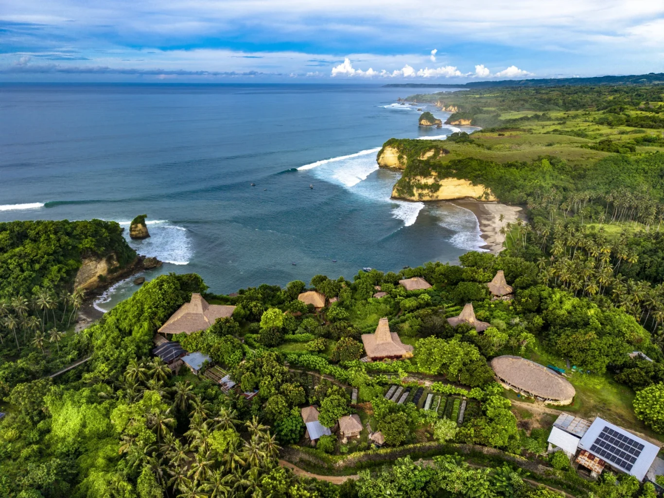 Aerial view of a vibrant green coastline with traditional thatched-roof buildings, palm trees, sandy beaches, and rocky cliffs along a deep blue ocean with white waves.