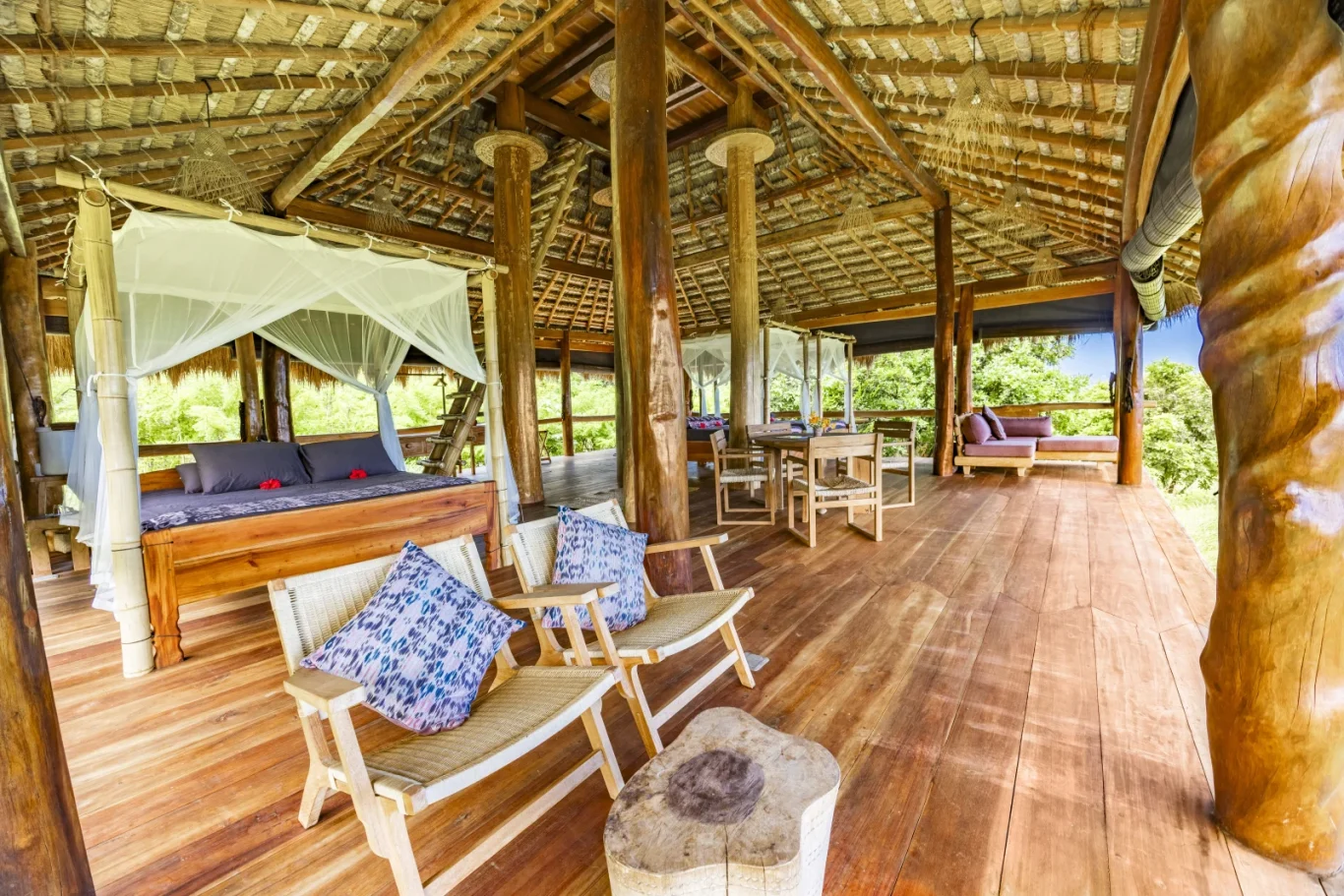 A spacious open-air room features a wooden bed with a mosquito net, woven chairs, and a dining set, all under a thatched roof, with green trees.