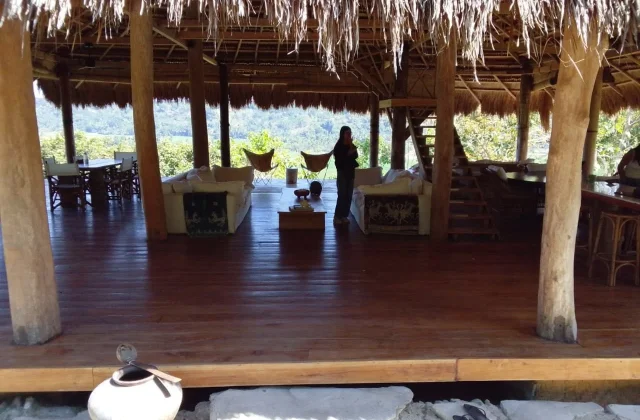 A person stands inside a large open-air, thatched-roof building with wooden floors and pillars, overlooking a lush green landscape.