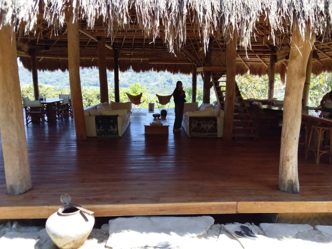 A person stands inside a large open-air, thatched-roof building with wooden floors and pillars, overlooking a lush green landscape.