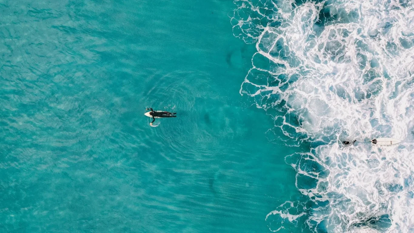 An aerial view shows two surfers in bright turquoise ocean water, one paddling calmly and the other near a foamy white breaking wave.