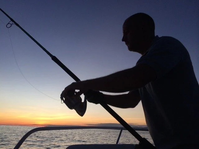 A man is silhouetted on a boat, holding a fishing rod and reeling it in against a sunset sky over the ocean.