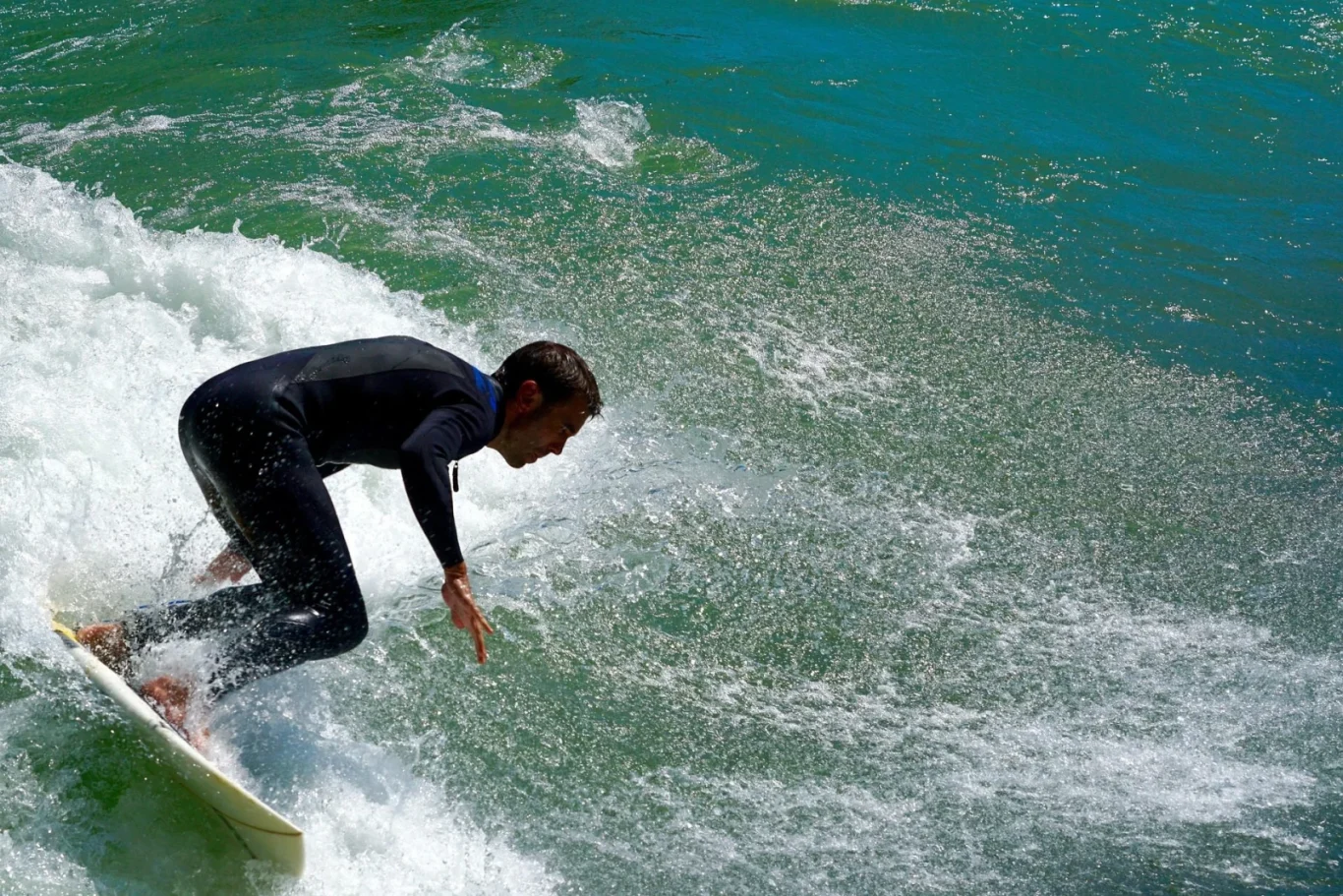 A man in a black wetsuit with blue accents surfs on a white board amidst turquoise water, white foam, and spray.