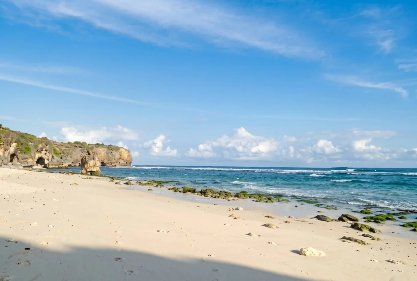 A wide sandy beach with footprints stretches towards a blue ocean with white waves, backed by a large rocky cliff under a partly cloudy sky.