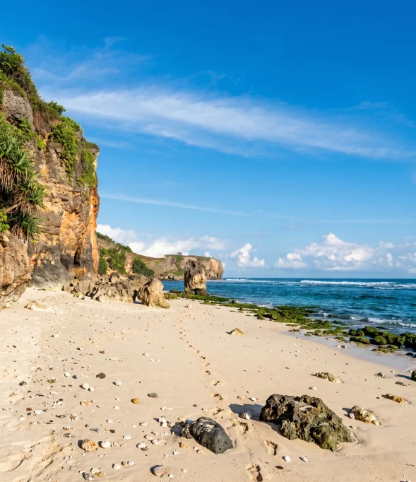 A wide, empty white sand beach stretches along a blue ocean with gentle waves, backed by green-topped rocky cliffs under a bright blue sky with wispy clouds.