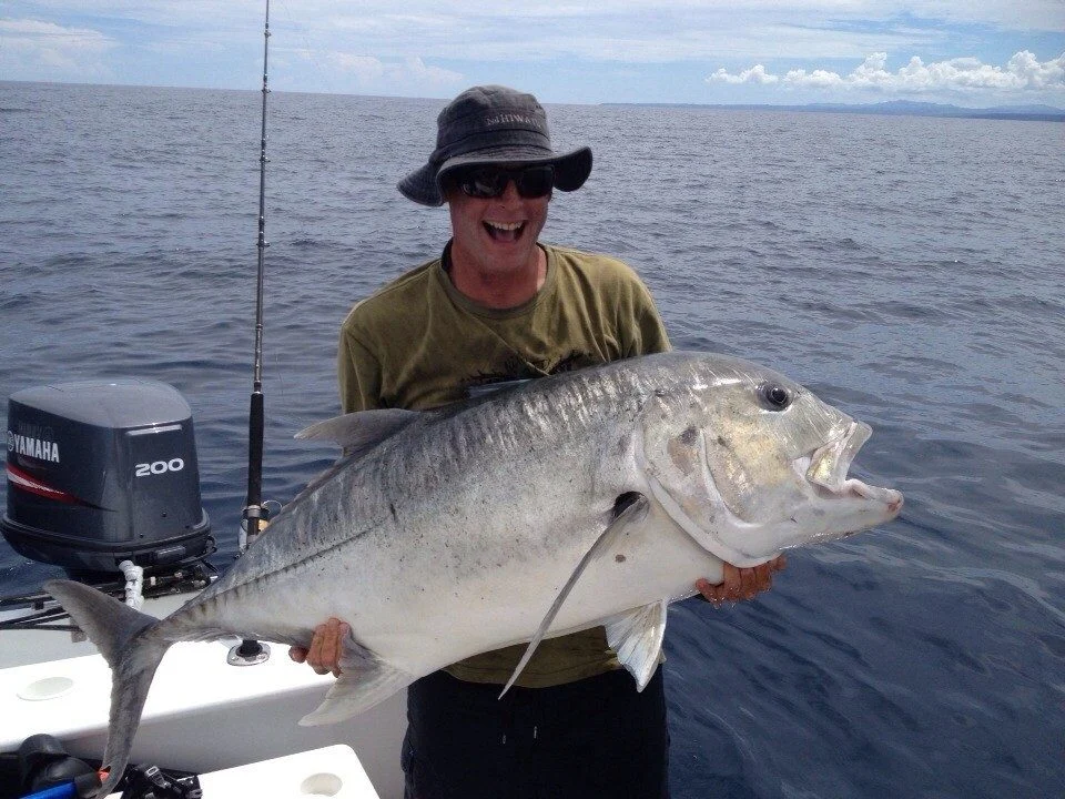 A smiling man in a hat and sunglasses holds a large silver-grey fish on a boat, with dark blue ocean and a partly cloudy sky in the background.