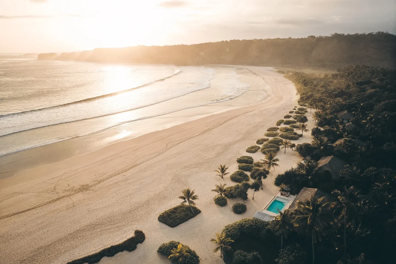 Aerial view of a golden sandy beach with gentle waves, lush green jungle, villas, and a blue pool, bathed in warm, hazy sunlight.