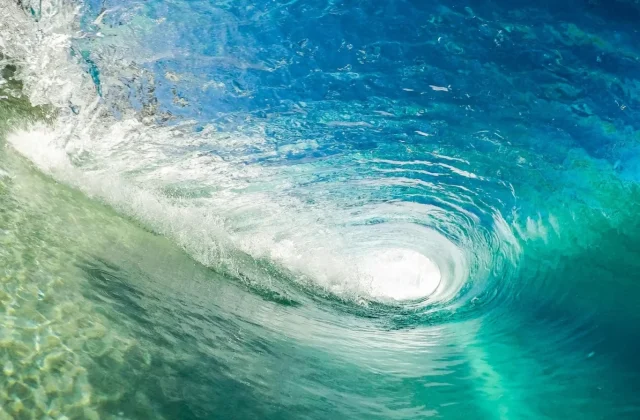 A clear blue and turquoise ocean wave curls into a barrel, with white foam and spray visible at the top and bright light shining through the opening.