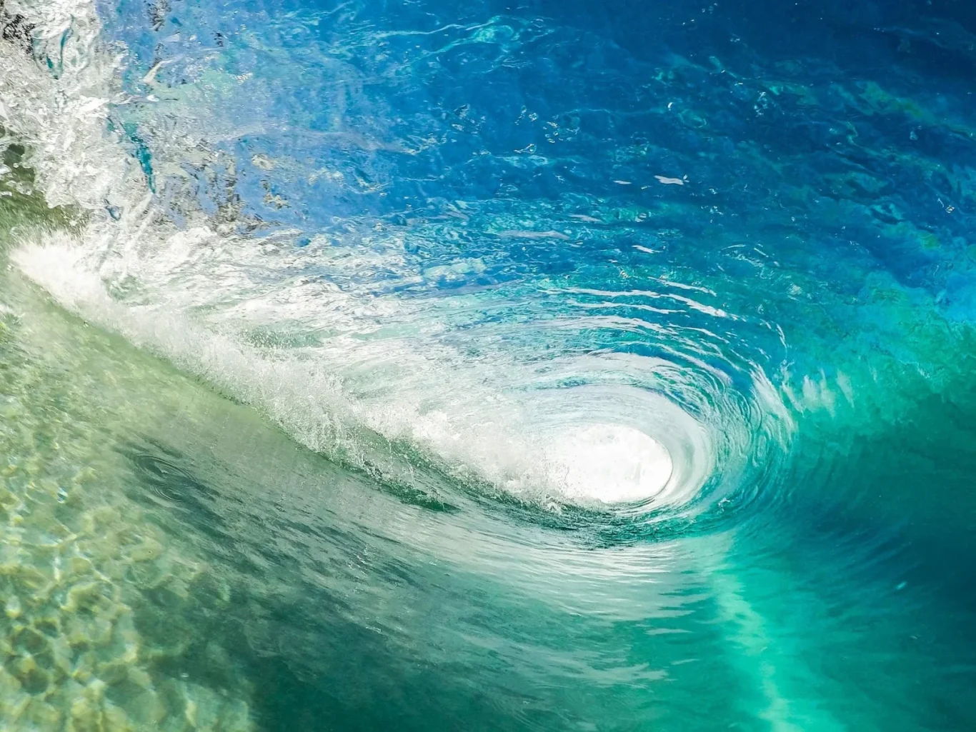 A clear blue and turquoise ocean wave curls into a barrel, with white foam and spray visible at the top and bright light shining through the opening.