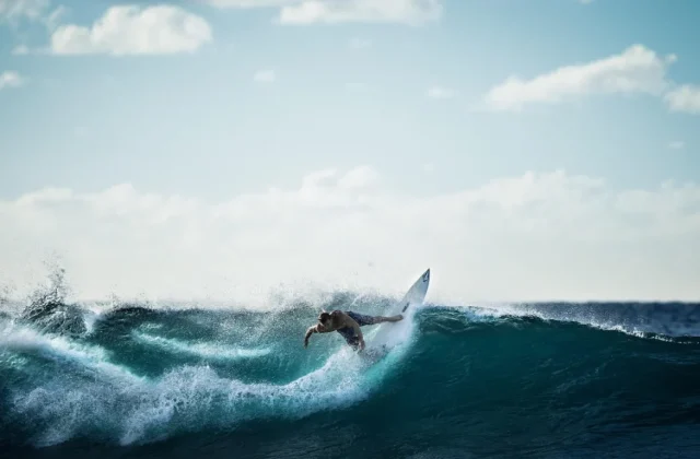 A male surfer on a white board carves a deep blue-green wave, creating white spray under a bright, partly cloudy sky.