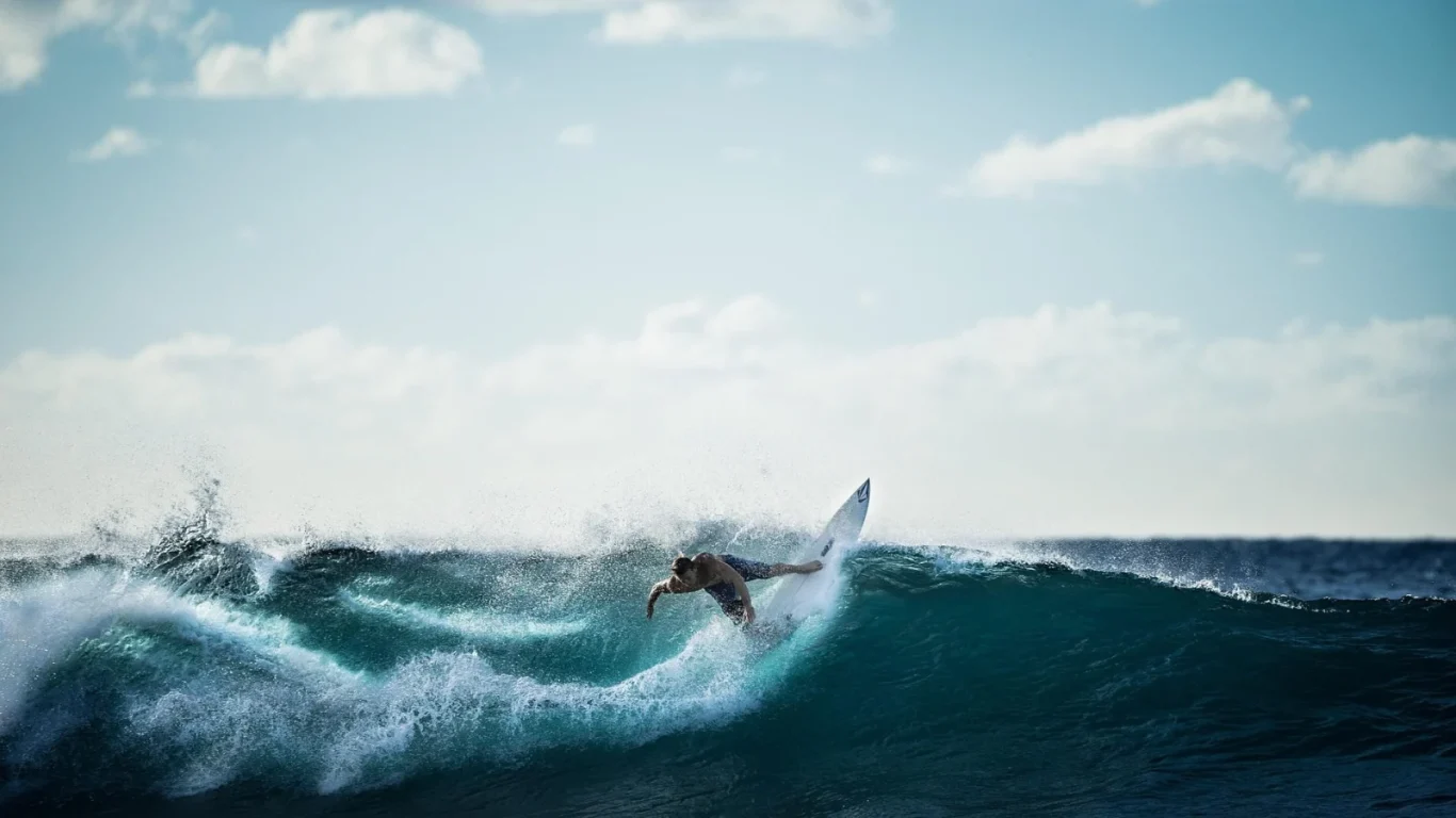 A male surfer on a white board carves a deep blue-green wave, creating white spray under a bright, partly cloudy sky.