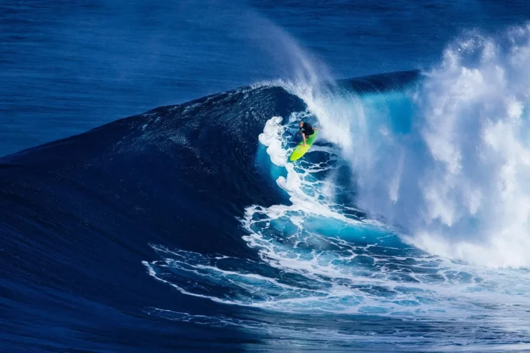 A surfer on a bright yellow-green board rides a large, deep blue ocean wave that is breaking with white foam and spray.