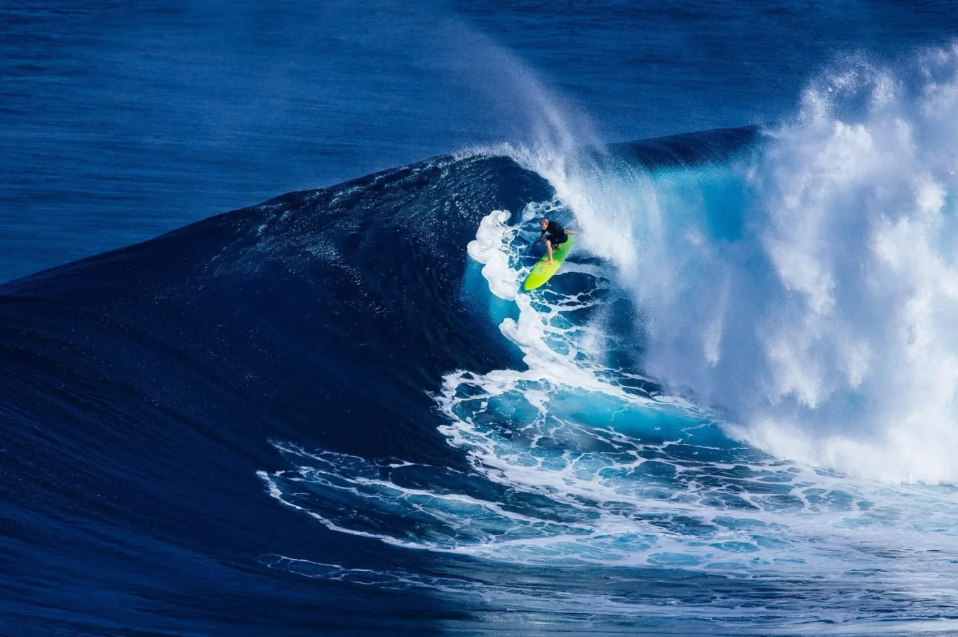 A surfer on a bright yellow-green board rides a large, deep blue ocean wave that is breaking with white foam and spray.