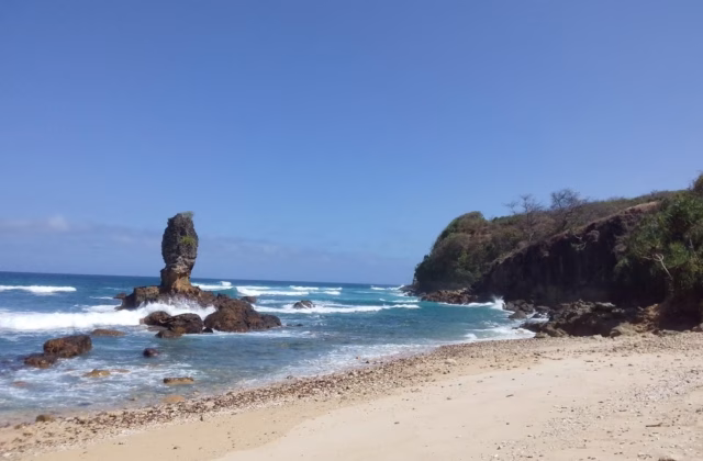 A prominent dark rock formation rises from the turquoise ocean with white waves, next to a sandy beach and a green cliff under a blue sky.