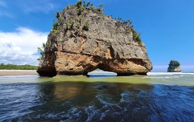 A large, brown and grey rock formation with an archway stands in the green-blue ocean water, with a sandy beach and green trees under a bright blue sky.