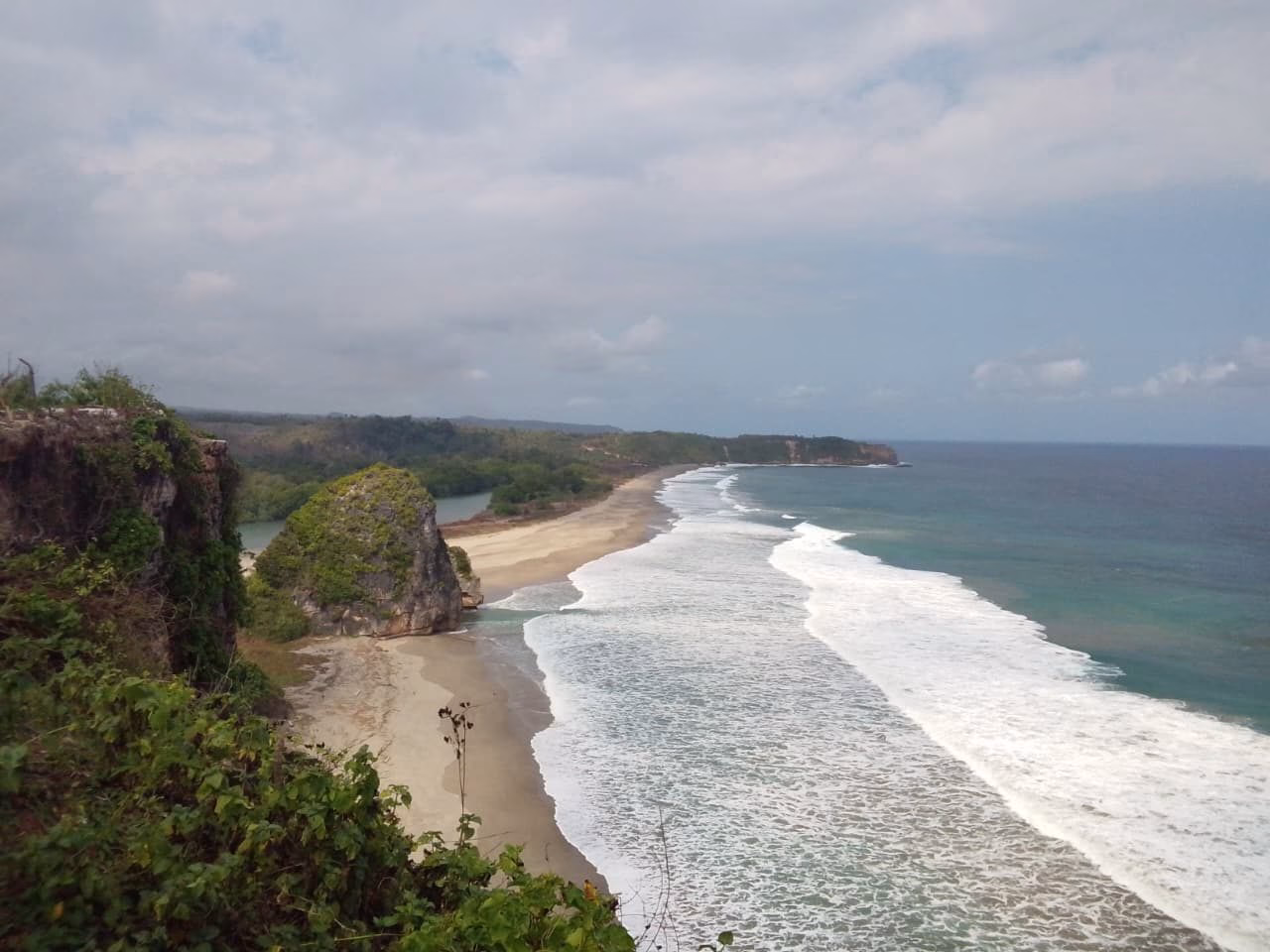 A wide coastal view shows a sandy beach with breaking white waves, a large mossy rock, and green cliffs under a partly cloudy sky.