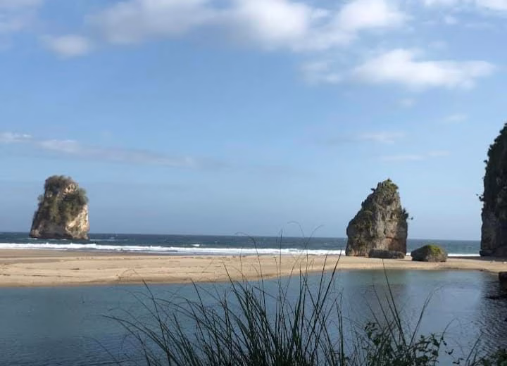 Two large, vegetated rock formations stand in the ocean near a sandy beach and a calm blue lagoon with tall grass in the foreground.