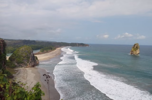 A wide coastal view shows a long sandy beach with white waves crashing, a large green-topped rock on the left, and a sea stack in the turquoise ocean under a cloudy sky.