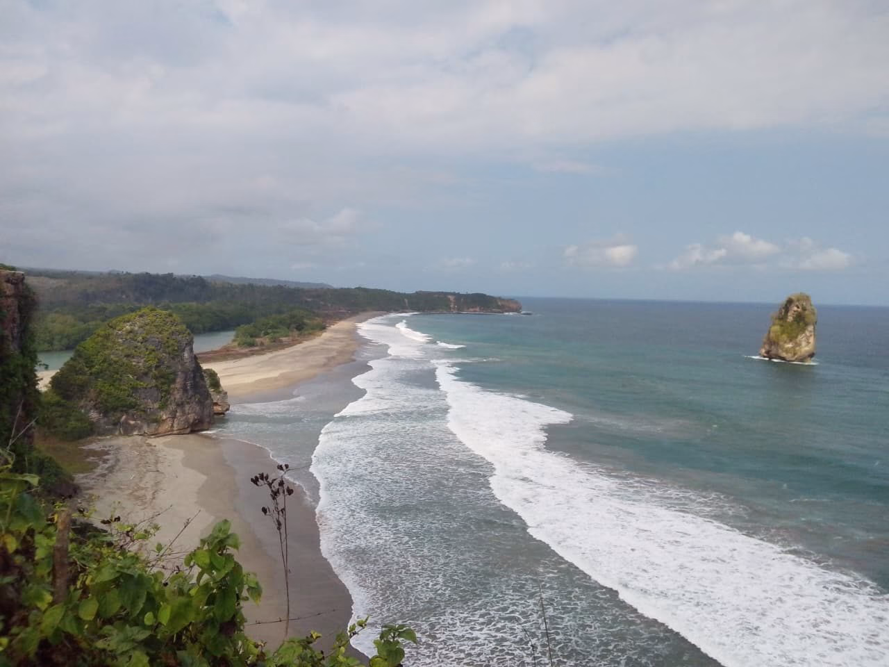 A wide coastal view shows a long sandy beach with white waves crashing, a large green-topped rock on the left, and a sea stack in the turquoise ocean under a cloudy sky.