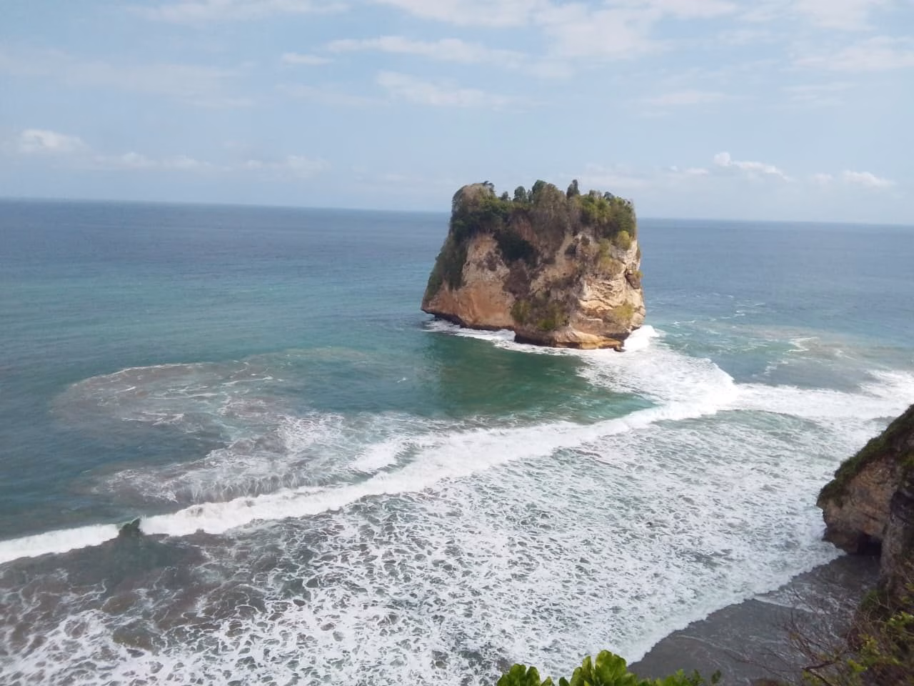 A large, tree-covered sea stack stands in a turquoise ocean with white waves crashing around it under a partly cloudy blue sky.