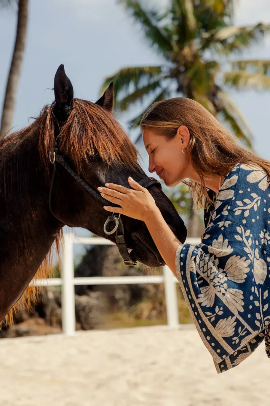 A woman with long brown hair gently touches the dark brown horse's face, smiling softly outdoors on sandy ground with palm trees.