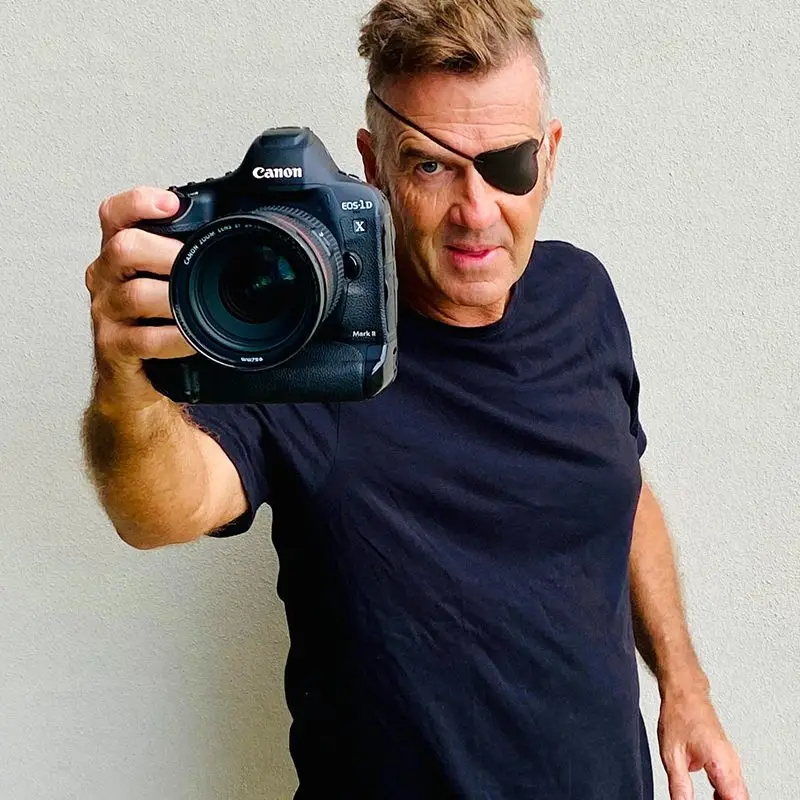 A man with an eye patch over his right eye holds a black Canon EOS-1D X Mark II camera with a lens pointed forward, against a light wall.