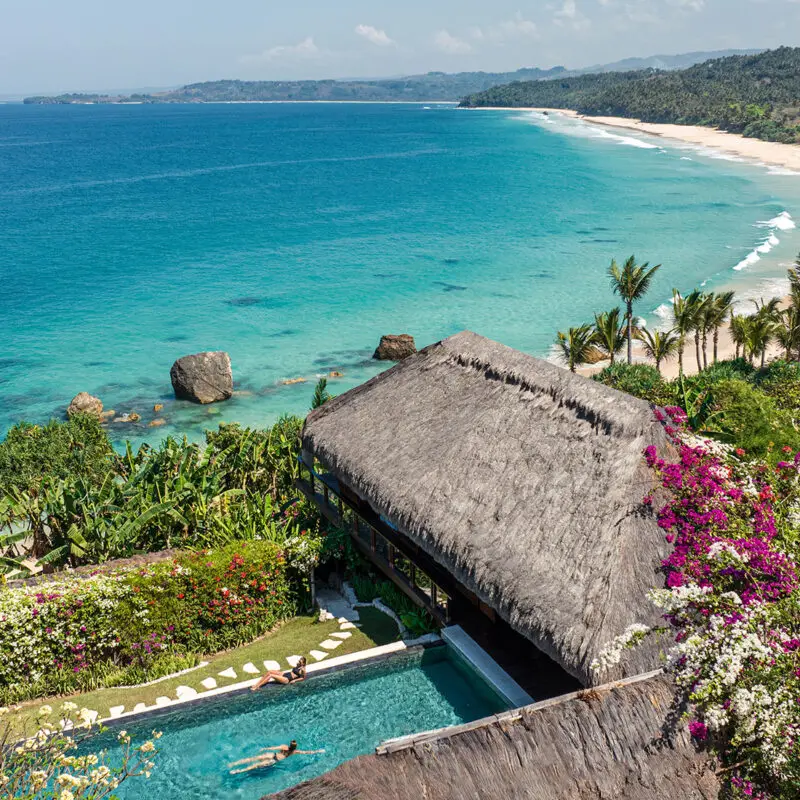 A thatched-roof villa, a private pool with two women, and lush flowers overlook a turquoise ocean and sandy beach with palm trees.