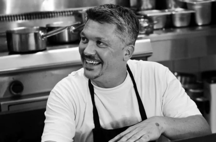 Numa Muller, a smiling man with graying hair and a goatee, wears a white t-shirt and dark apron in a black and white kitchen portrait.