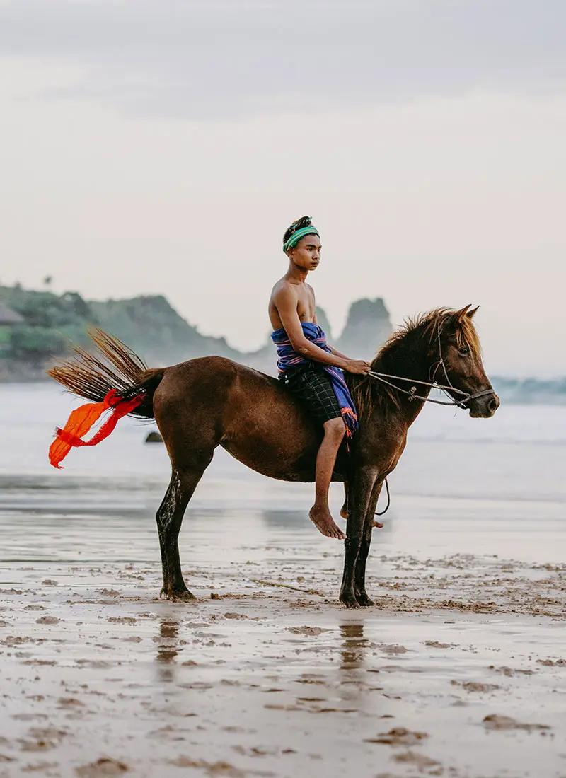A young man in a blue sarong and green head wrap rides a brown horse bareback on a wet sandy beach under an overcast sky.