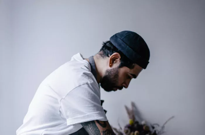 Chef Robert Mendoza, a bearded man in a black beanie and white shirt, looks down while arranging dried botanicals against a light grey background.