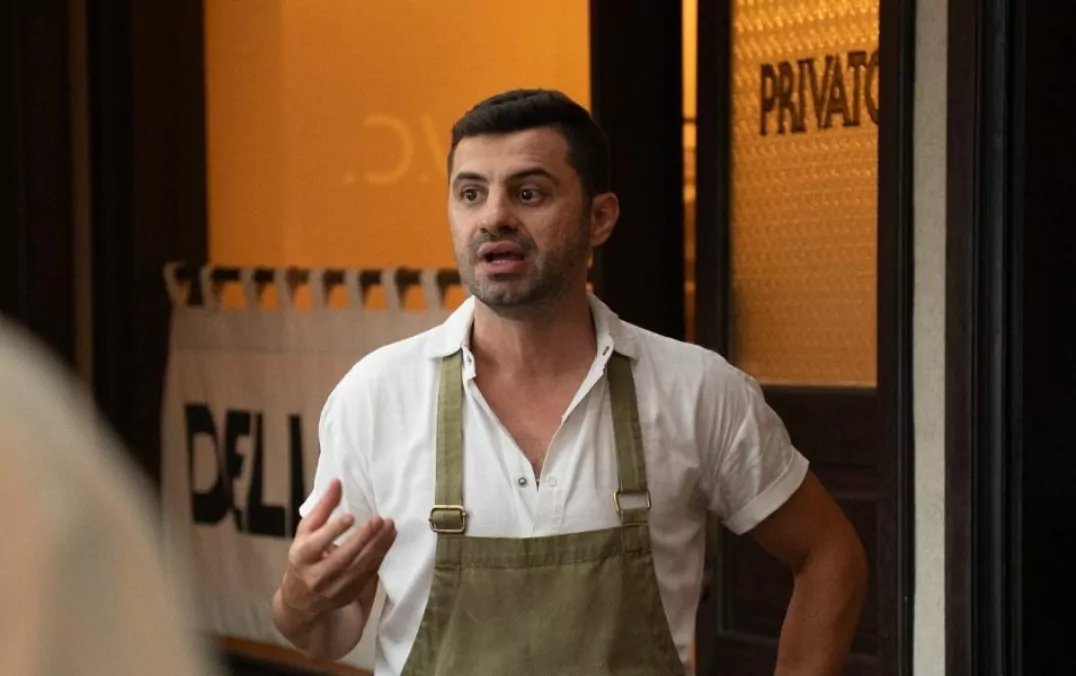 A man with dark hair and a beard, wearing a white shirt and green apron, speaks and gestures indoors against an orange wall.